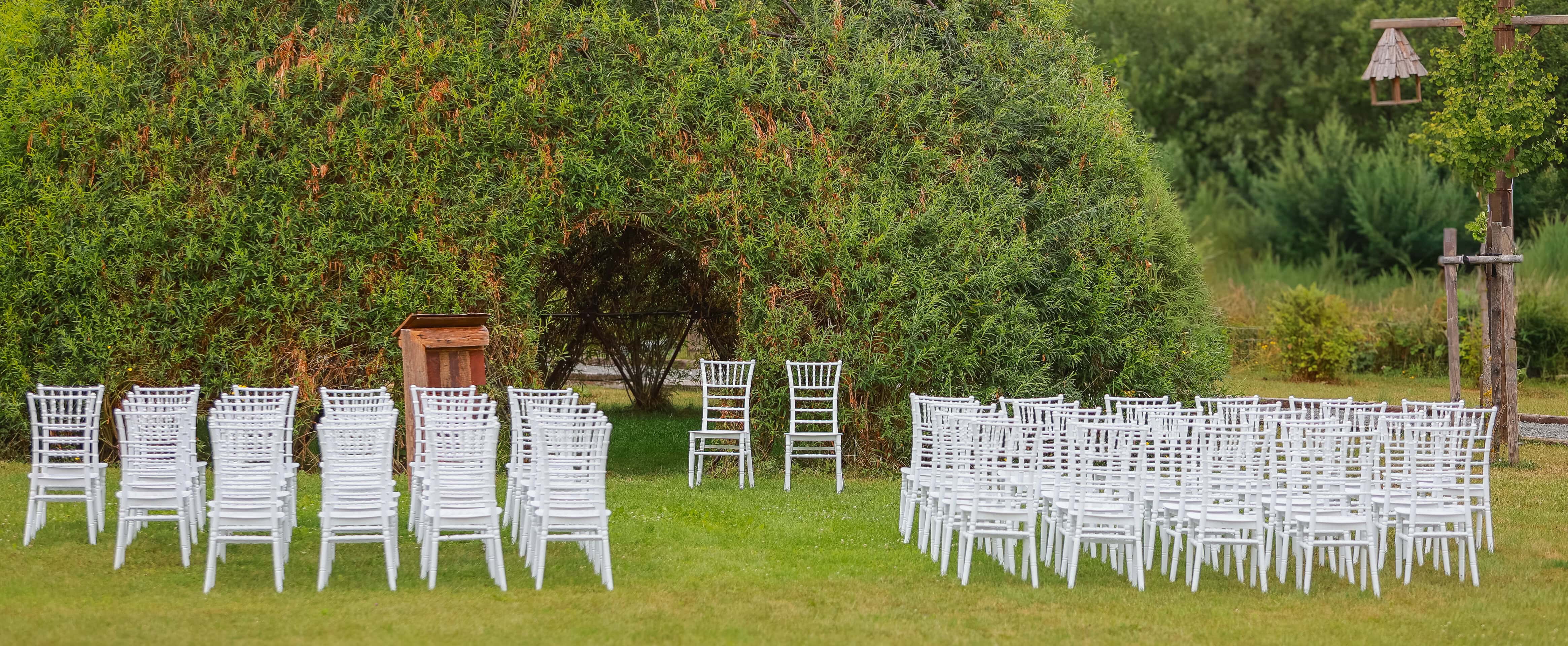 Aufgestellte weiße Stühle für eine freie Trauung im Grünen vor einer natürlichen Weidenkulisse.